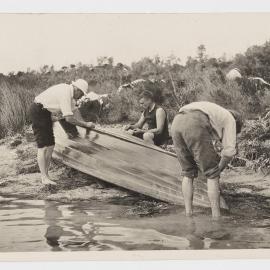 [Three men leaning over upturned row boat]