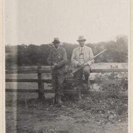 [Two men holding shotguns posing on fence]