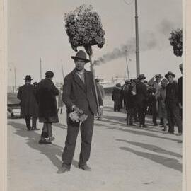 [Man wearing suit and hat holding arrangement of flowers on long wooden stick]