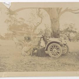 [Man and woman seated in front of motor car]
